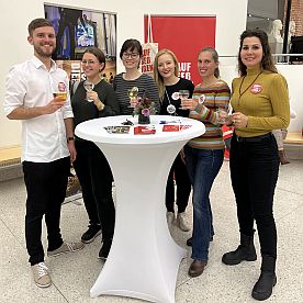 Einige der jungen Freunde stehen an einem Tisch im Museum und prosten den Besucher*innen zu.