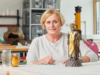 Simone Hänisch sitzt an einem Tisch in der Restaurierungswerkstatt. Eine kleine Skulptur aus dem 19. Jahrhundert steht vor ihr.