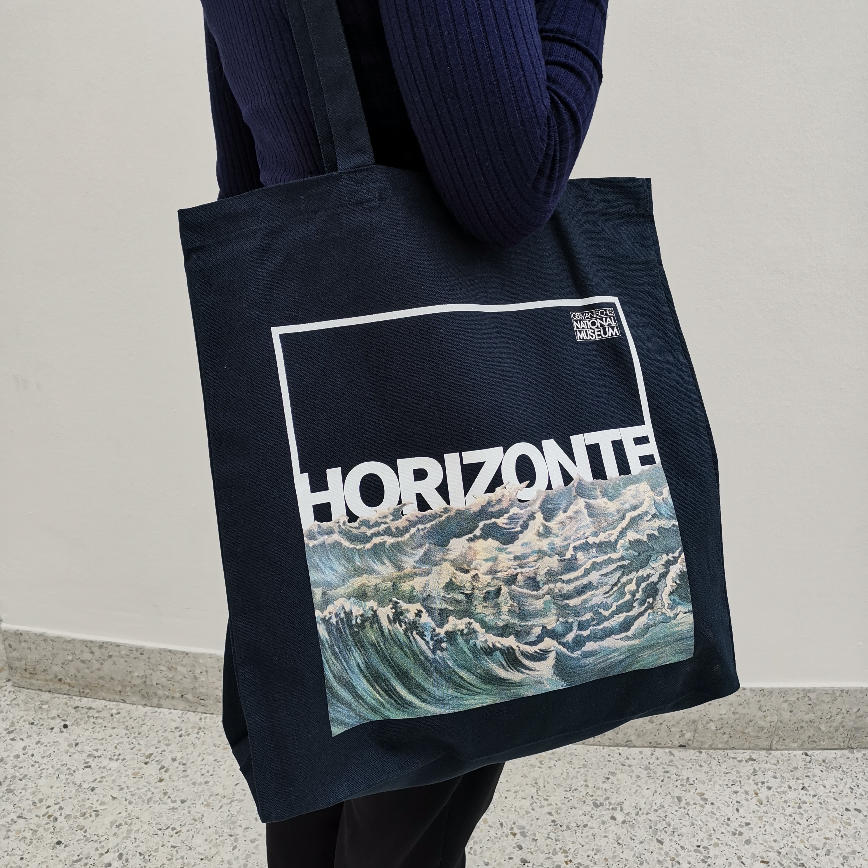 Eine blaue Stofftasche mit langen Henkeln und dem Aufdruck des Motives der Ausstellung, ein brausendes Meer und den Titel "Horizonte".